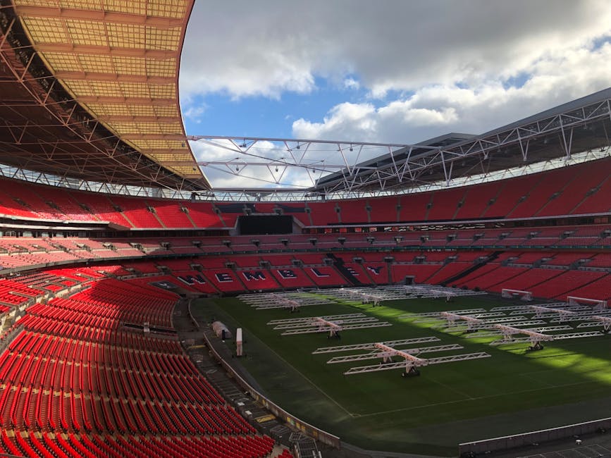 Inside an empty stadium with red seating arrangements, the view captures a large central field being prepared for an event, with several white staging platforms and wheeled equipment arranged on the grass. The seating area is vast, with tiered sections surrounding the pitch, and the word 'WEMBLEY' visible spelled out in black seats on one side. The stadium roof structure, which is partially open, reveals a bright blue sky with scattered clouds, allowing natural light to illuminate the scene. There are no people present in the image, emphasizing the staging and setup process for a potential concert or sports event. This setting highlights the logistical aspects of home relocation or event transport, illustrating how large venues like Wembley Stadium are prepared for different functions, with equipment and staging materials carefully arranged on the field. Brent Movers efficiently handles such operations, offering comprehensive removals and furniture transport services that often involve staging and loading processes in large-scale venues.