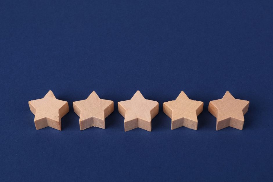 Five light-colored wooden star-shaped objects arranged in a horizontal line on a plain blue background, with the front star slightly closer to the camera. The objects are smooth and evenly shaped, with sharp edges defining each star. The blue surface appears flat, and the lighting creates soft shadows underneath each star, emphasizing their three-dimensional form. The arrangement suggests a simplified representation, possibly used for visual communication of ratings or quality. In the context of house removals or moving services, these star-shaped objects could symbolise quality ratings or customer feedback, aligning with services offered by Brent Movers for home relocation and furniture transport. The image is neutral and professional, suitable for use on a webpage providing advice and rates for small flat removals in Willesden Green, London.