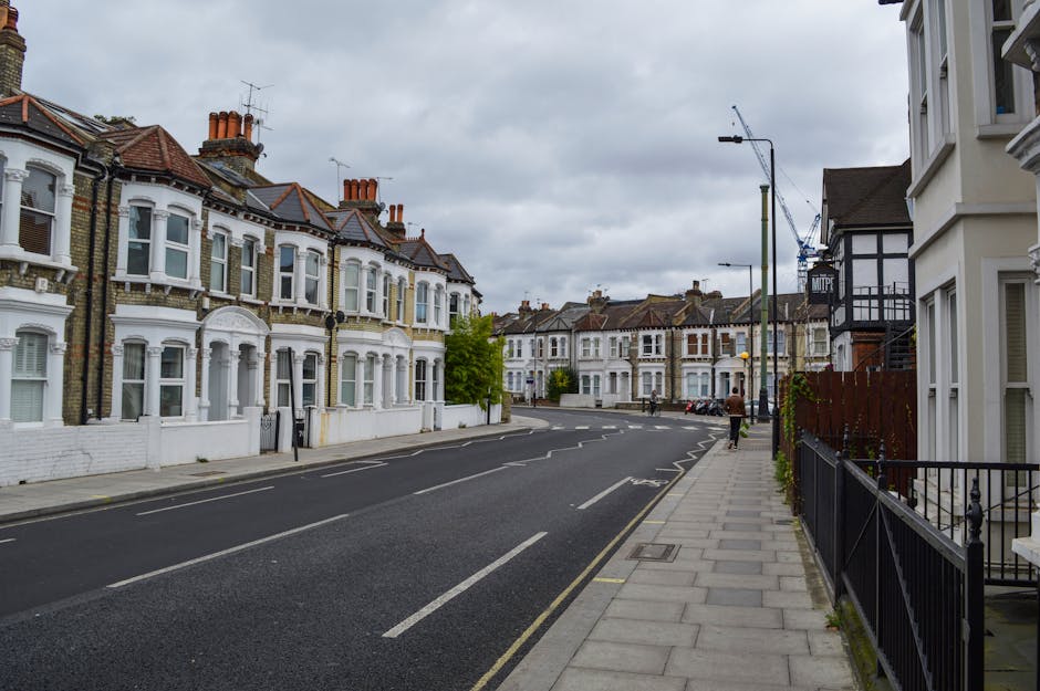 A quiet residential street in Willesden Green with a row of white Victorian-style terraced houses featuring bay windows and brick chimneys on the left side. The pavement runs alongside these houses, with a black metal railing in front of the immediate property, and a few small plants visible on the right. The road has gently curved lanes with white road markings and no vehicles parked along the side. Overcast skies cast diffuse light over the scene. In the distance, there are more houses, a crane, and street lamps. This setting is typical for house removals and furniture transport in the area, where careful planning is required for home relocation logistics, especially when loading furniture and boxes onto the nearby street or vehicle loading area. Brent Movers, specializing in removals, operates in this neighbourhood, providing packing and moving services suitable for small flat relocations in Willesden Green.